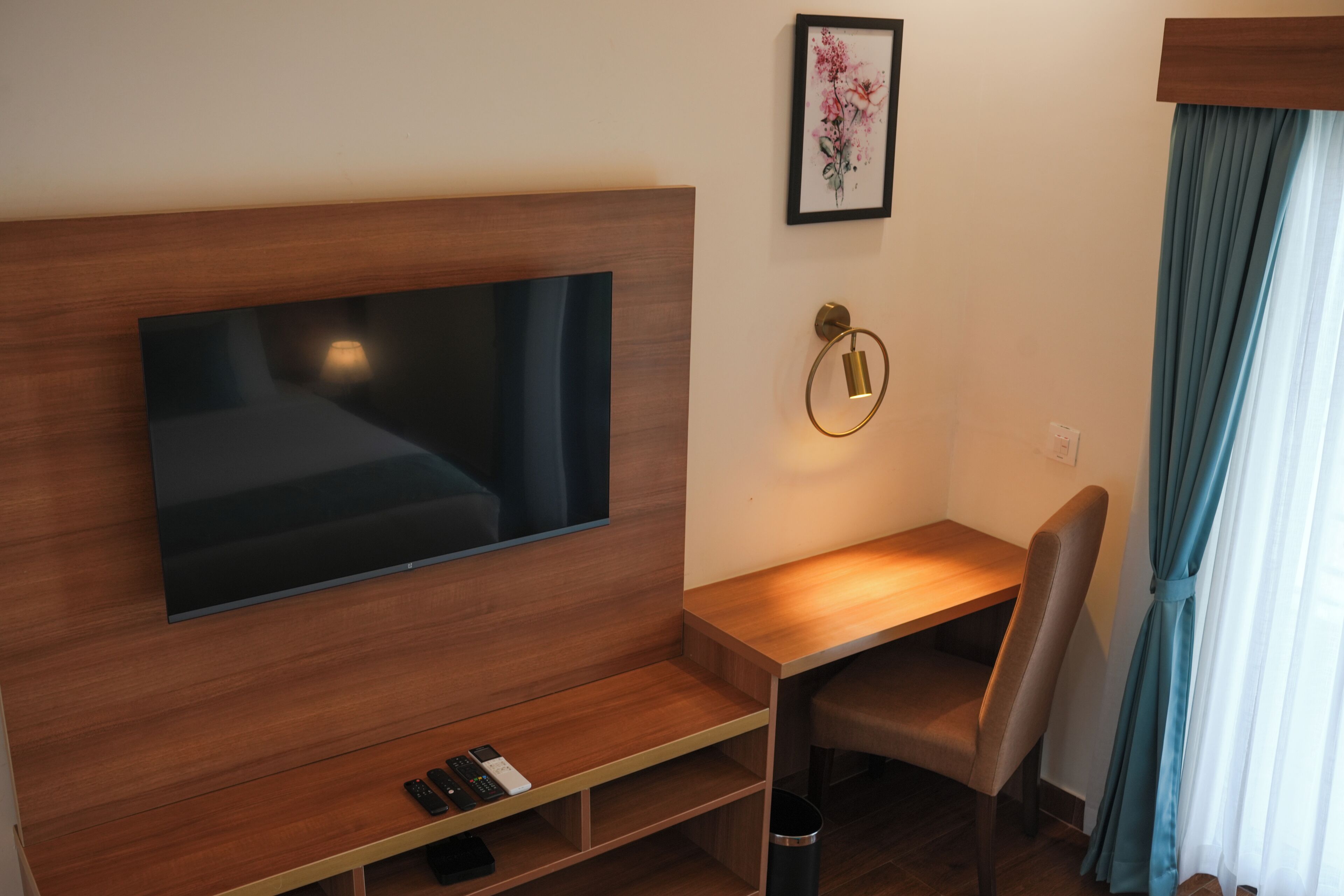 Habitación de hotel con un televisor de pantalla plana grande montado en una pared de madera, un escritorio con una silla, un cuadro enmarcado en la pared y una ventana con cortinas.