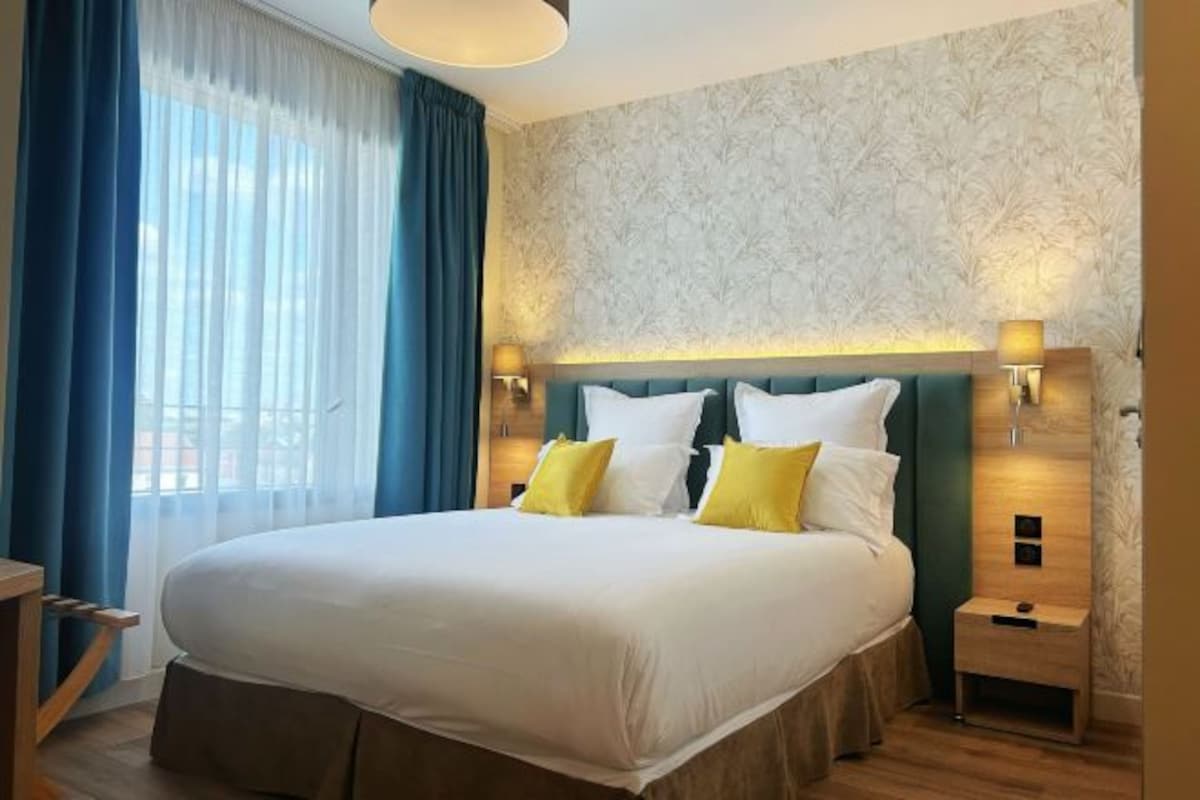 A hotel room with a large bed, a nightstand, a lamp, and a window with curtains.