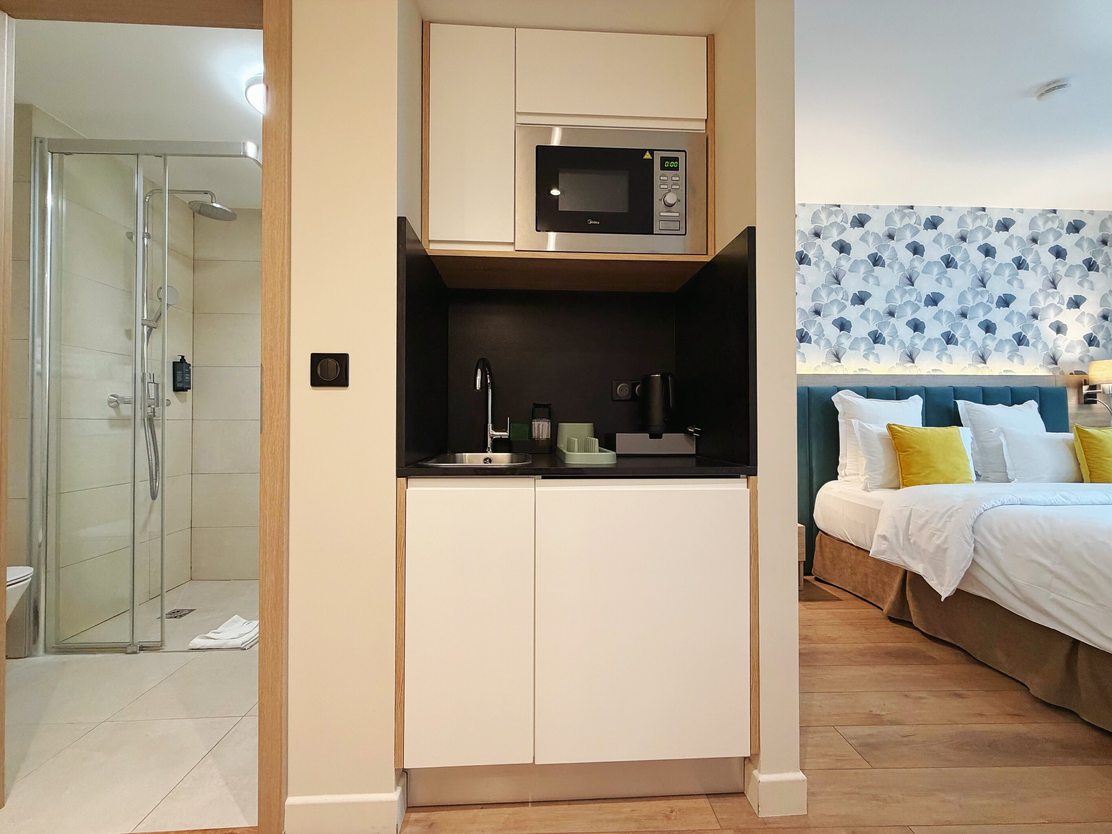 A compact hotel room with a kitchenette, a shower area, and a bed with a patterned headboard.