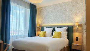 A hotel room with a large bed, two bedside tables, a nightstand, a window with blue curtains, and a wall with floral wallpaper. - Hôtel Oden Paris Ivry (Ivry-sur-Seine)