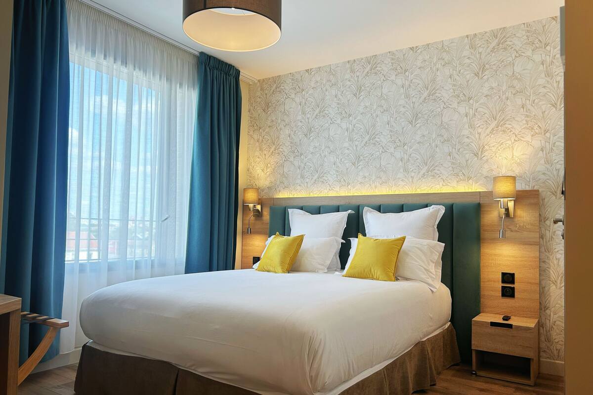Ein Hotelzimmer mit einem großen Bett, zwei Nachttischen, einem Fenster mit blauen Vorhängen und einer Wand mit floralem Wandpapier.