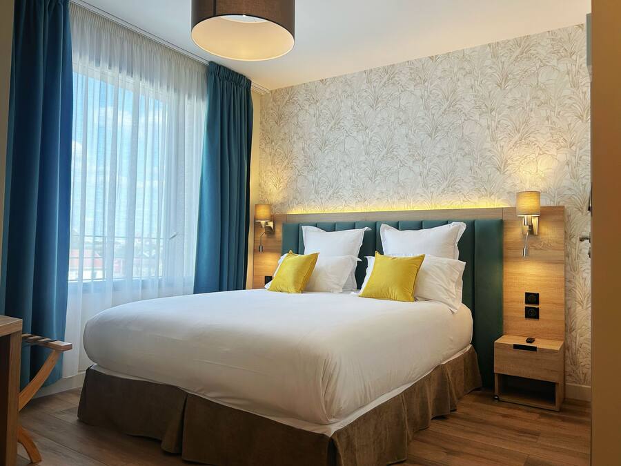 Una habitación de hotel con una cama grande, dos mesitas de noche, un buró, una ventana con cortinas azules y una pared con estampado floral.