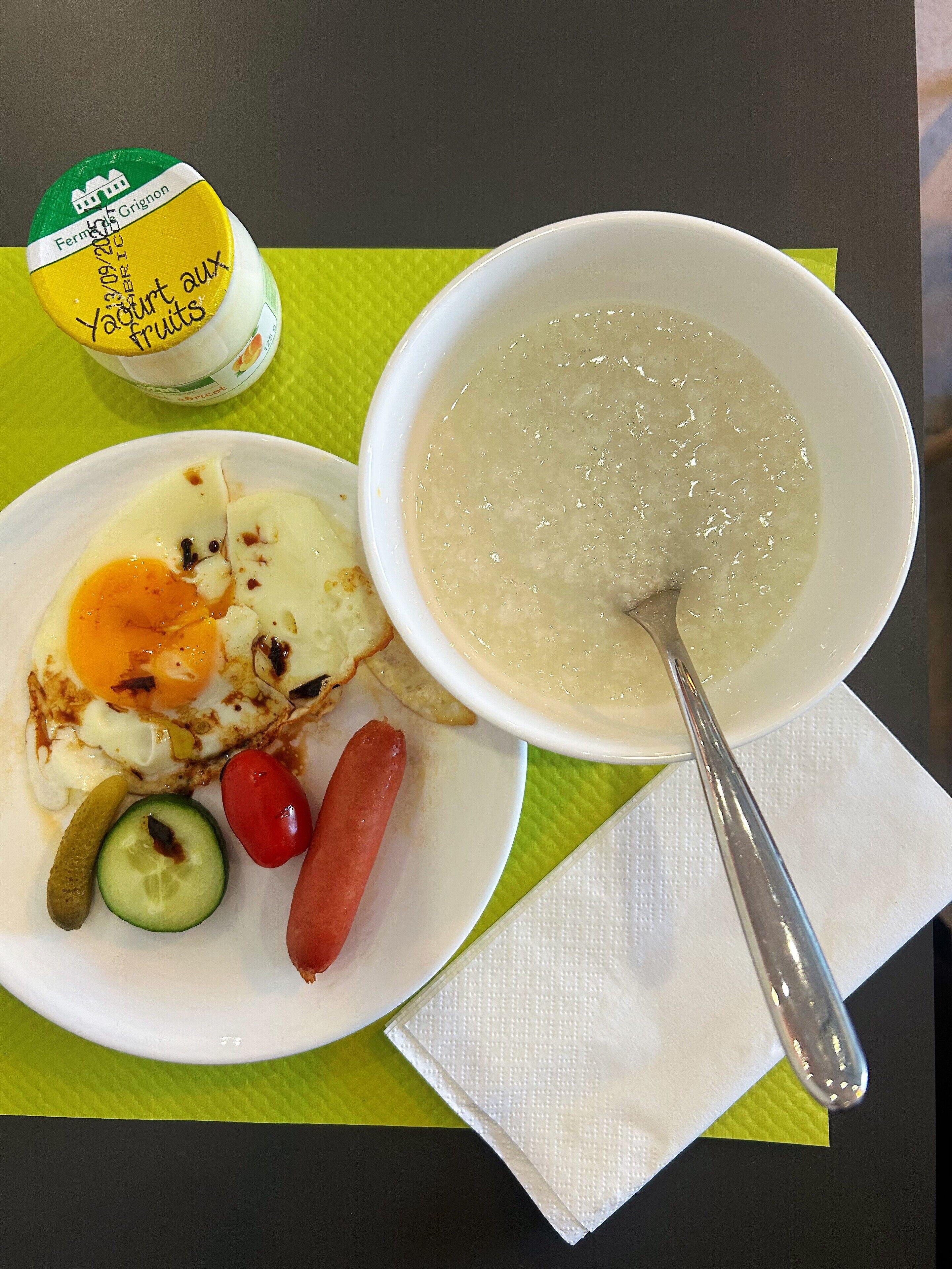 A meal consisting of a bowl of soup, a plate with eggs, a sausage, and a pickle.