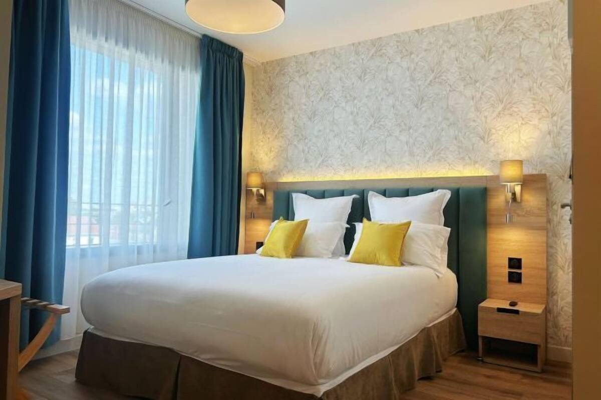 Ein Hotelzimmer mit einem großen Bett, einem Nachttisch, einer Lampe und einem Fenster mit Vorhängen.