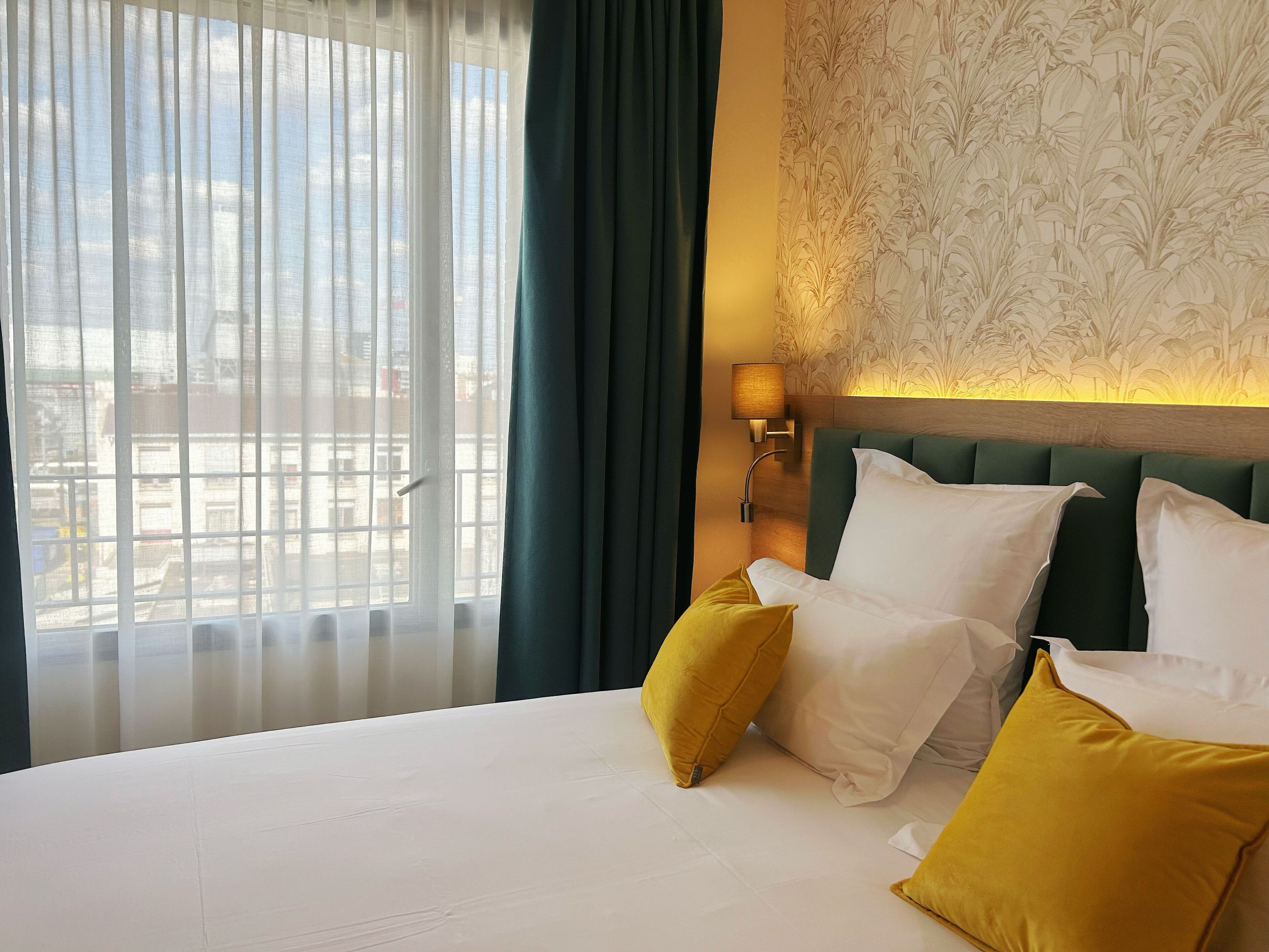 Quarto de hotel com cama grande, duas almofadas, mesa de cabeceira, luminária e vista para prédios através de cortinas finas.
