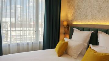 Quarto de hotel com cama grande, duas almofadas, mesa de cabeceira, luminária e vista para prédios através de cortinas finas.