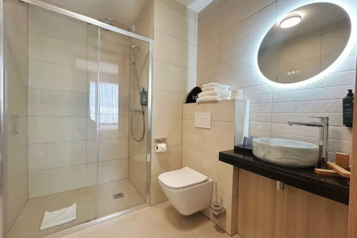 A modern bathroom with a glass-enclosed shower, a white toilet, a round mirror, and a black countertop with a sink.