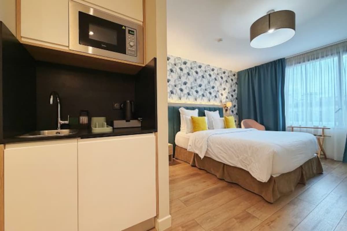 A compact hotel room with a kitchenette, a bed with white bedding, a blue patterned wall, and a large window with curtains.