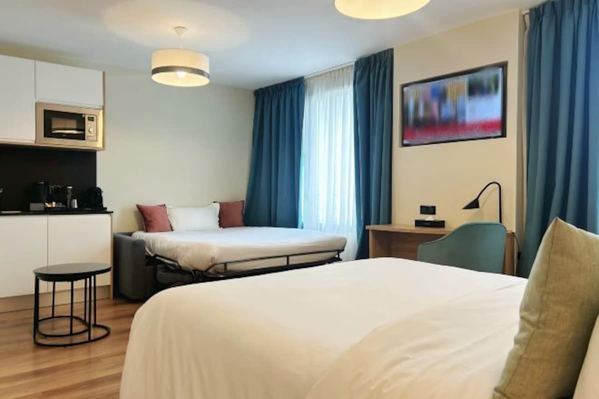 A hotel room with a bed, a desk, a chair, a small table, a microwave, and a painting on the wall.