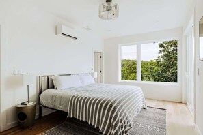 A bedroom with a bed, a window, an air conditioning unit, and a bedside table.