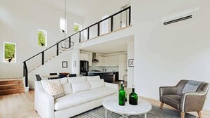 Un vaste espace de vie comprenant un canapé blanc, une table basse ronde et un fauteuil gris. Un escalier mène à un niveau supérieur où se trouve une cuisine, visible depuis le rez-de-chaussée.