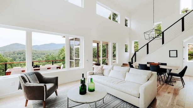 Un salon spacieux avec un canapé blanc, un fauteuil marron et un coin repas comprenant une table et des chaises. De grandes fenêtres offrent une vue sur l’extérieur.