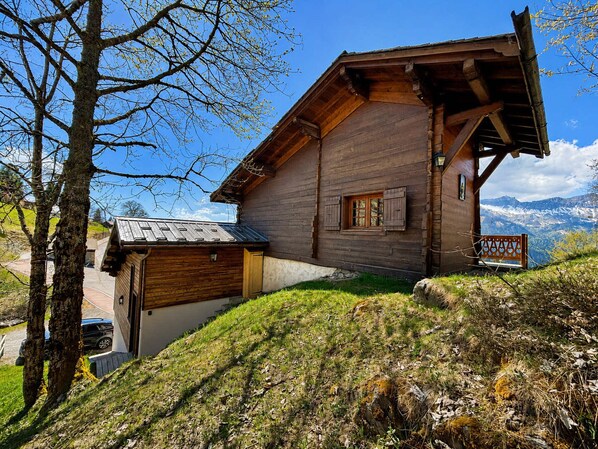 Una cabaña de madera con techo inclinado, un pequeño garaje adosado y una vista de montañas al fondo.