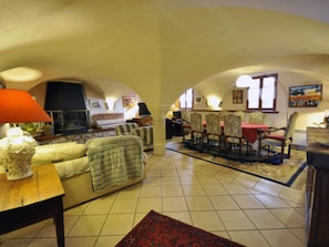 A cozy living room with a fireplace, a dining area with chairs, and a kitchen in the background.