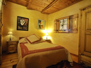 A bedroom with a wooden ceiling, a bed with a quilt, two bedside lamps, and a window with shutters.