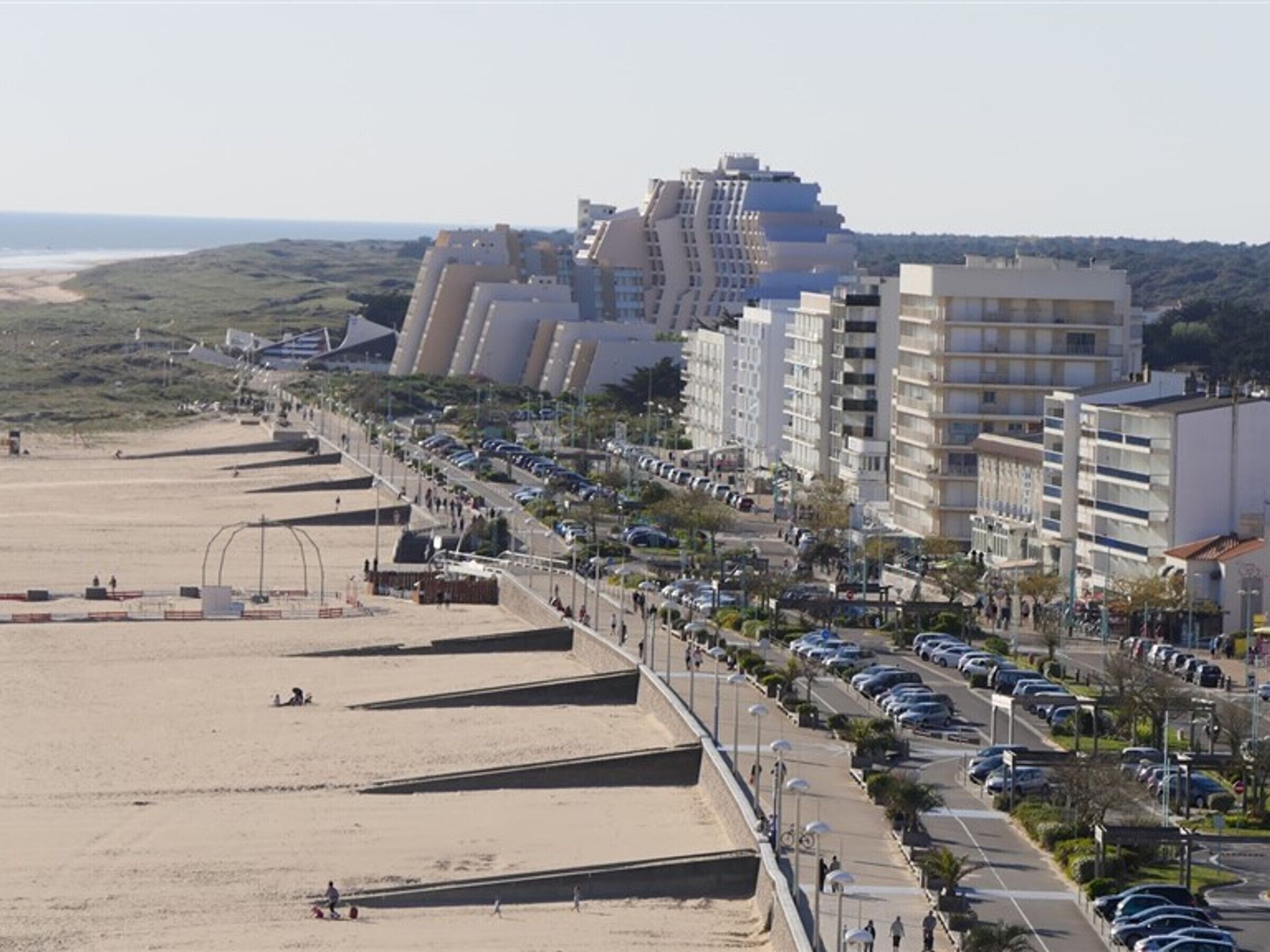 A coastal cityscape with a beach, modern buildings, and a parking area.