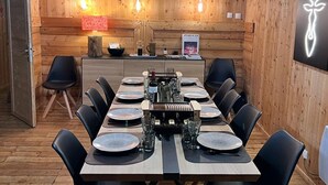 A wooden interior with a dining table set for four, chairs, a kitchen area, and a wall-mounted light fixture. - Spacious 6-Room Chalet for 10, Private Garden, Near Serre Chevalier Pistes (La Salle-les-Alpes)