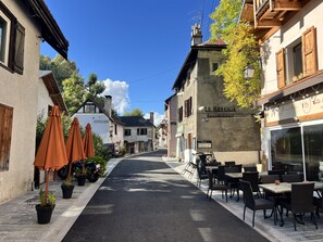 Een straat met terrasjes, een restaurant genaamd 'Le Refuge Restaurant', en traditionele Europese architectuur.