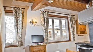 Een gezellige kamer met een houten plafond, een televisie op een houten kast, een tafel met een kleurrijke tafelkleed en een keukenhoek met een fornuis.