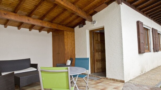 Een overdekt terrasgedeelte met houten plafond, een bank, een tafel met stoelen en een deur naar een binnenruimte.