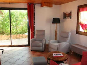Une pièce avec deux fauteuils gris, un plancher en bois, un rideau rouge, un lampadaire, une petite table avec un livre et une fenêtre donnant sur un paysage verdoyant.