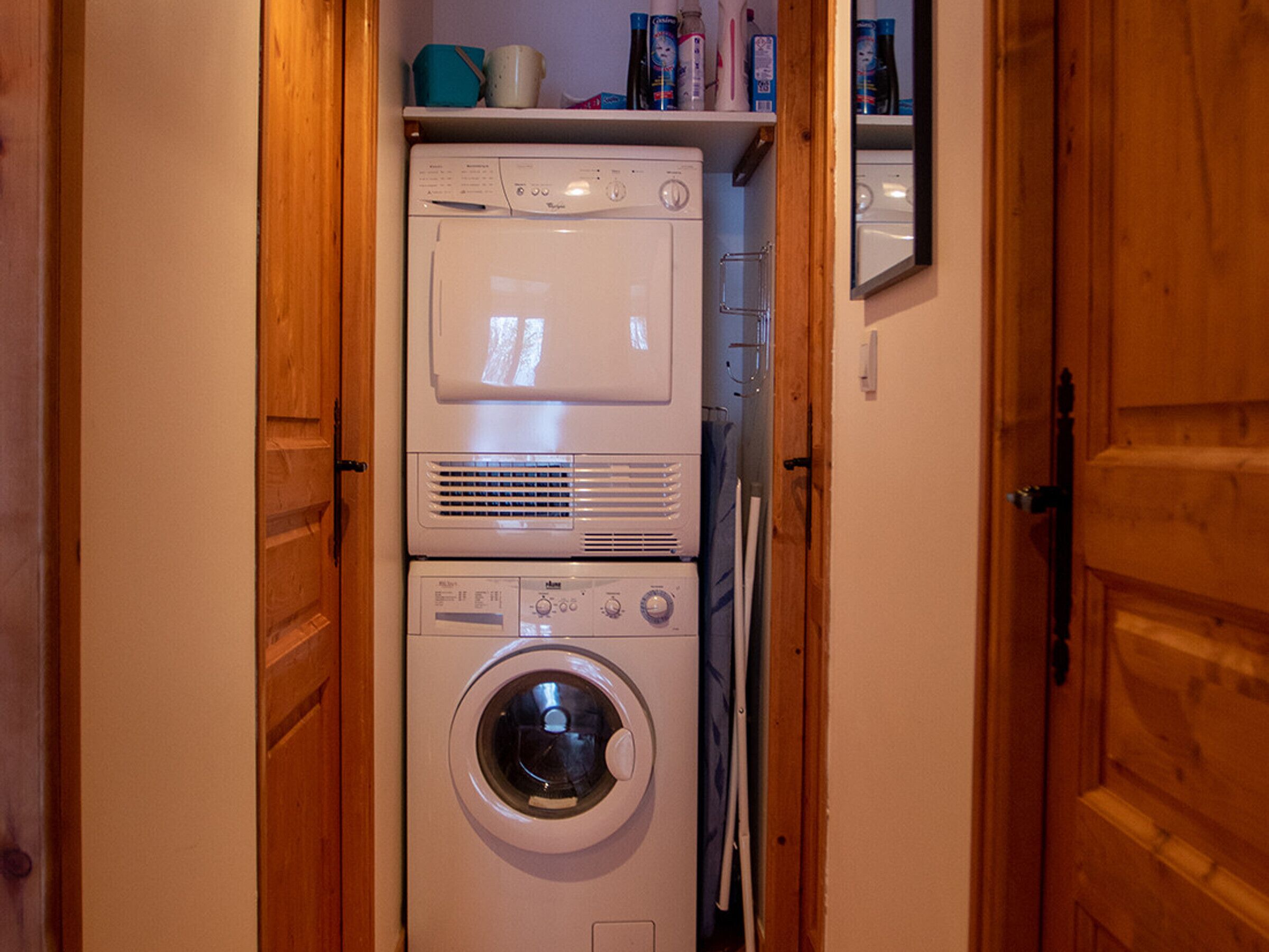 A laundry room with stacked washers and dryers, wooden doors, and a shelf with cleaning supplies.