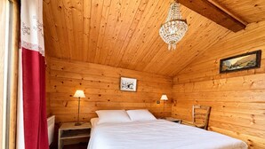 Un dormitorio de cabaña de madera con cama, dos lámparas de noche, una araña, un cuadro en la pared y una ventana con cortinas.