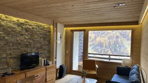 L’intérieur d’un chalet accueillant, avec un mur en pierre, des meubles en bois, un téléviseur et une grande fenêtre donnant sur la forêt.