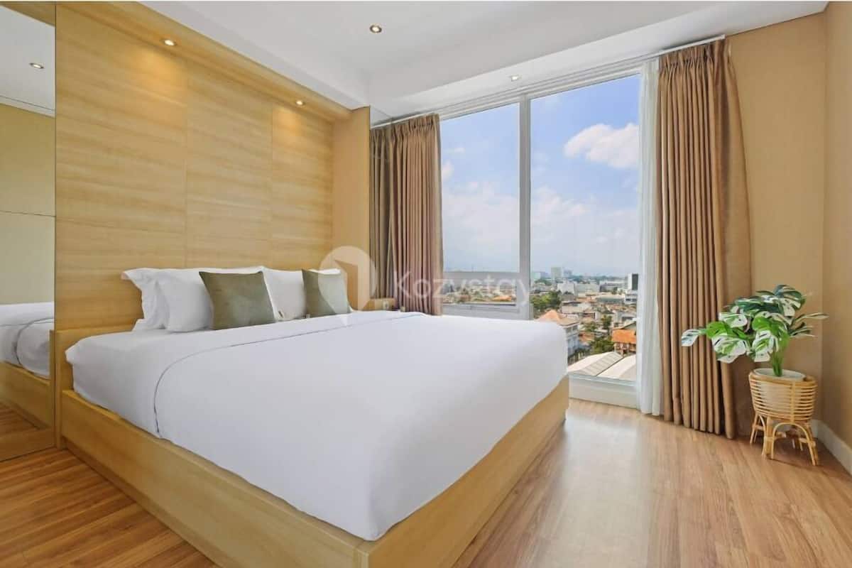 A modern bedroom with a large bed, wooden headboard, and a view of a cityscape through large windows.