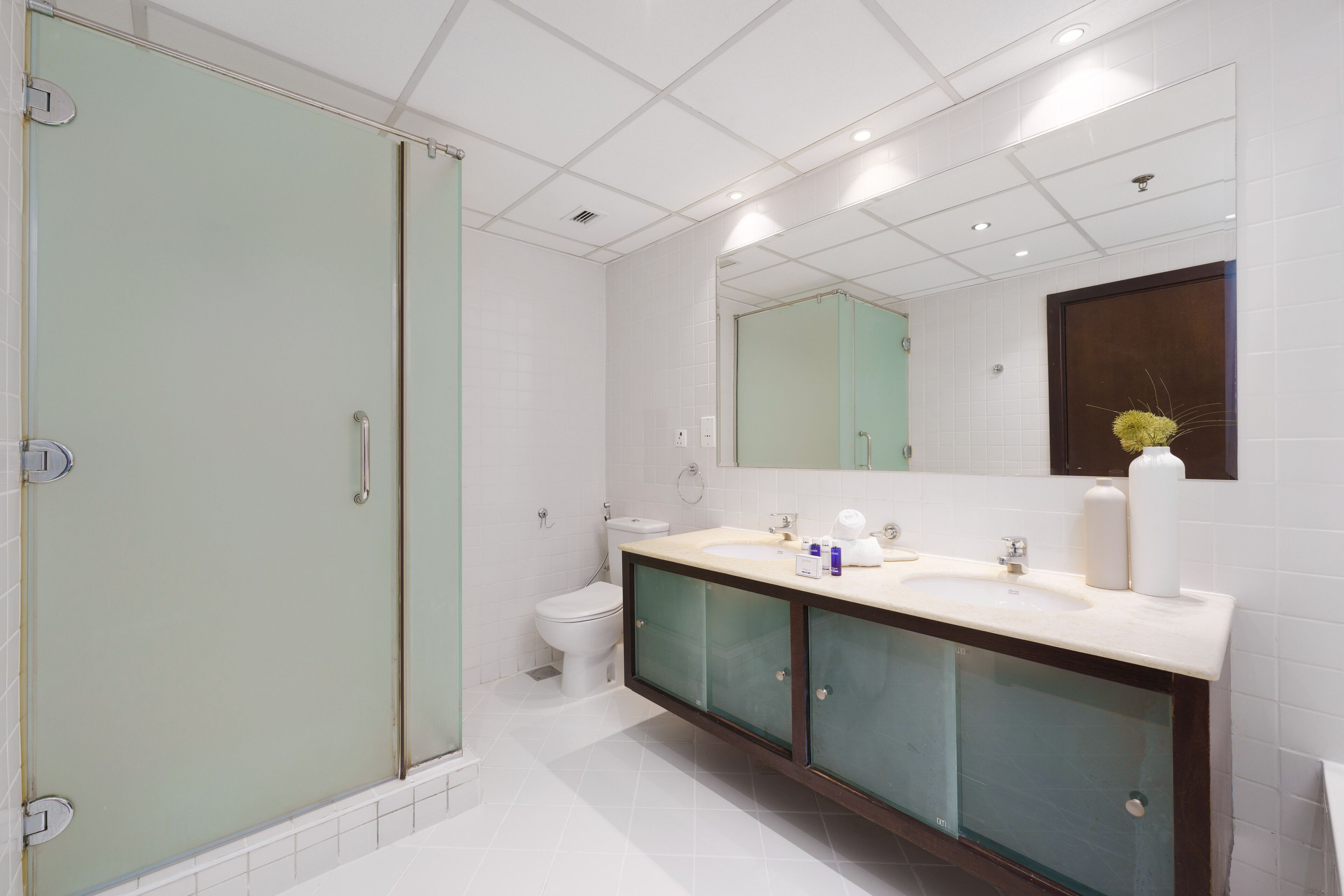Un baño moderno con cabina de ducha de vidrio, un inodoro blanco y un tocador con espejo y dos lavamanos.
