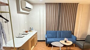 A modern hotel room with a wooden floor, a blue sofa, a small round table with a vase, a kitchen area with a marble countertop, and a hanging air conditioner. - d'primahotel Jemursari Surabaya (Surabaya)