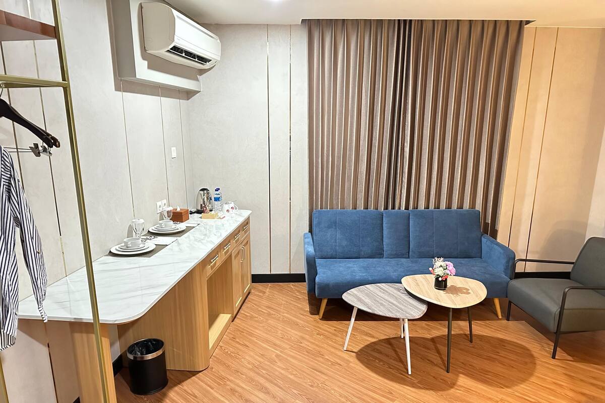 Ein modernes Hotelzimmer mit Holzboden, einem blauen Sofa, einem kleinen runden Tisch mit einer Vase, einer Küchenzeile mit Marmorarbeitsplatte und einer hängenden Klimaanlage.