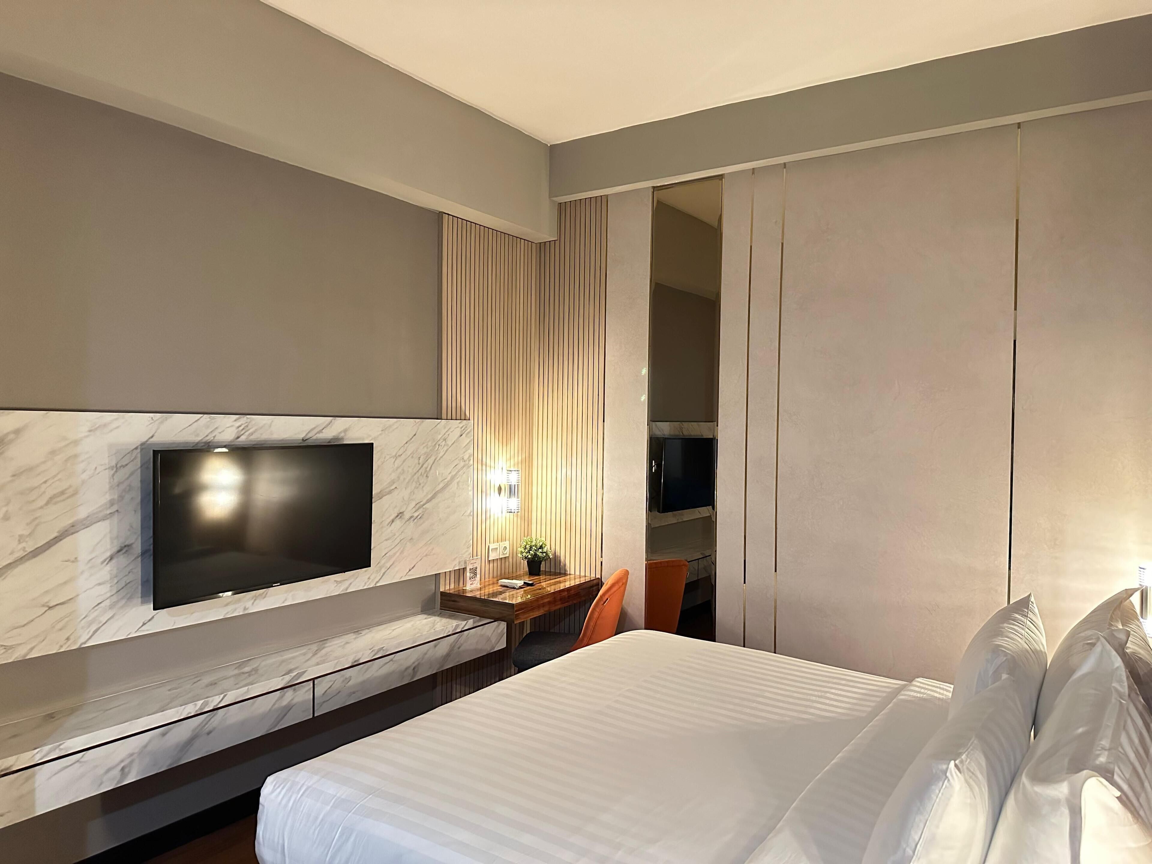 A modern hotel room with a large bed, a flat-screen TV mounted on a marble wall, a wooden nightstand, and a built-in wardrobe.