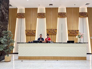 A reception desk with two staff members, a decorative wall with vertical panels, and a potted plant.