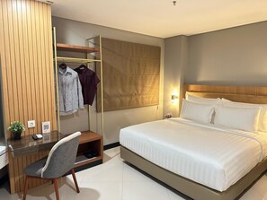 A hotel room with a bed, a desk with a chair, a wardrobe, and a small plant.