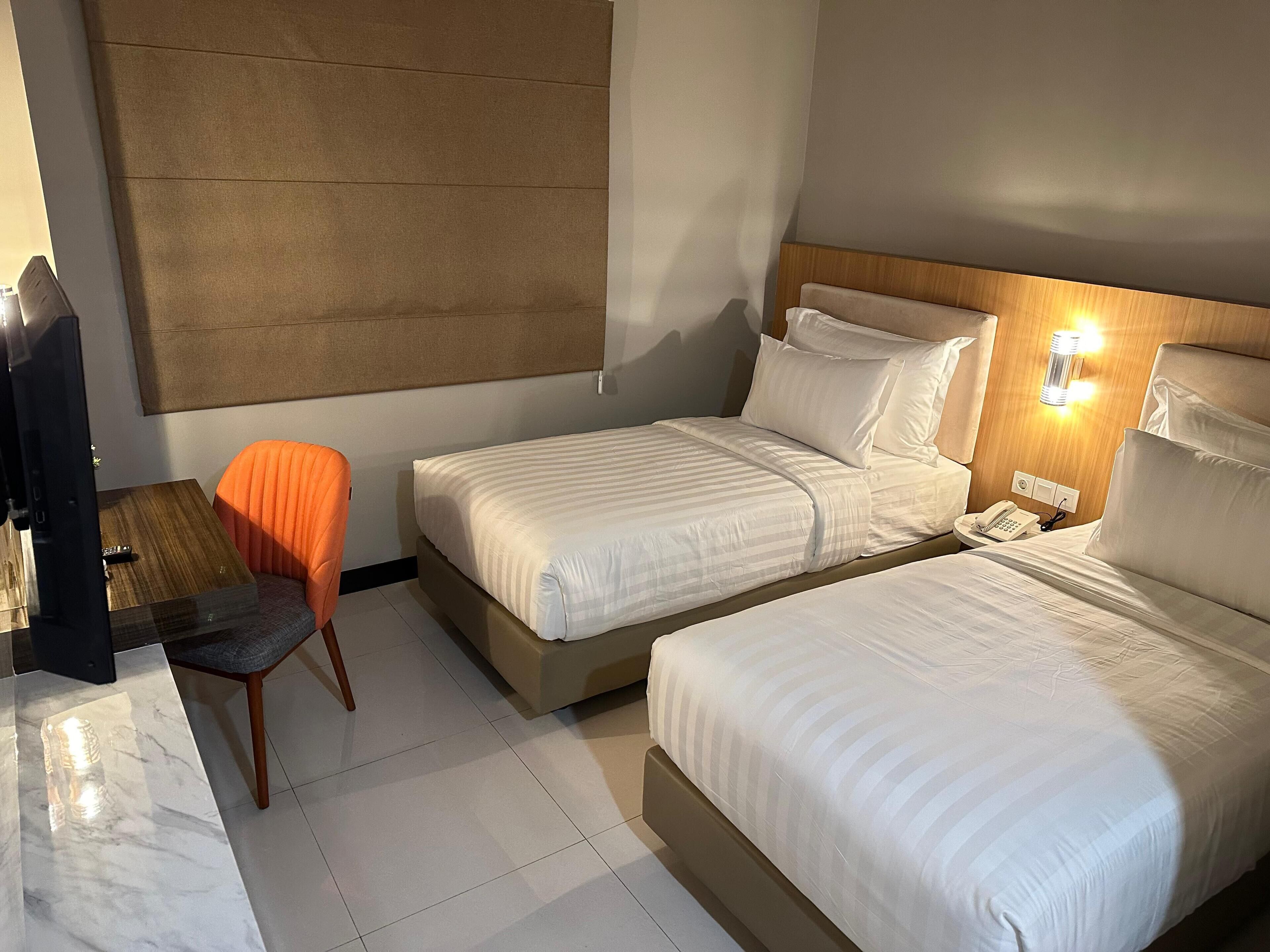 A hotel room with two beds, a small table with a remote control, an orange chair, and a wooden headboard.