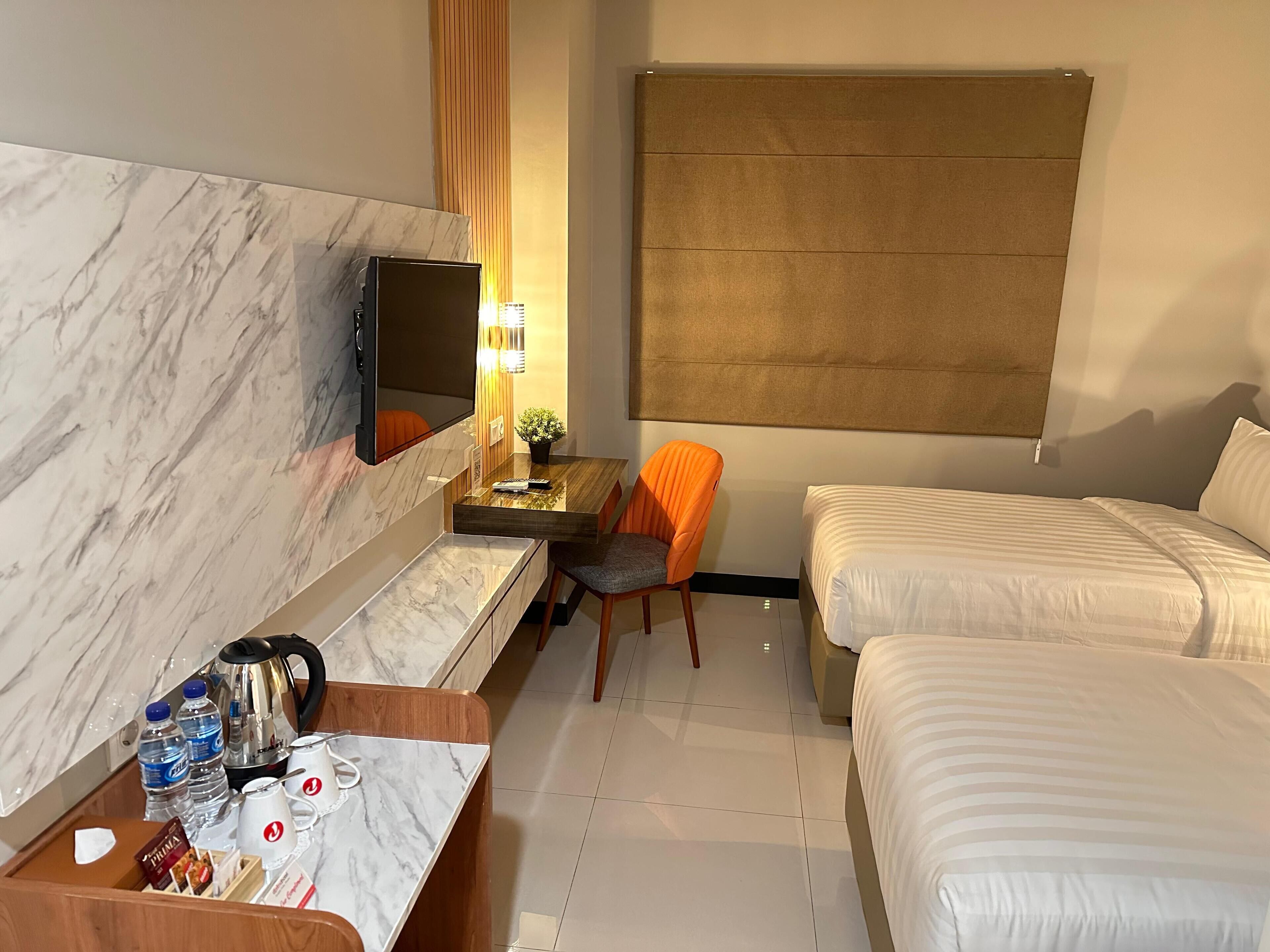 A hotel room with a marble headboard, a television, a desk with a plant, two beds, and a window with a brown blind.