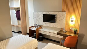 A hotel room with two beds, a desk with a flat-screen TV, a wooden panel wall, and a small potted plant. - d'primahotel Jemursari Surabaya (Surabaya)