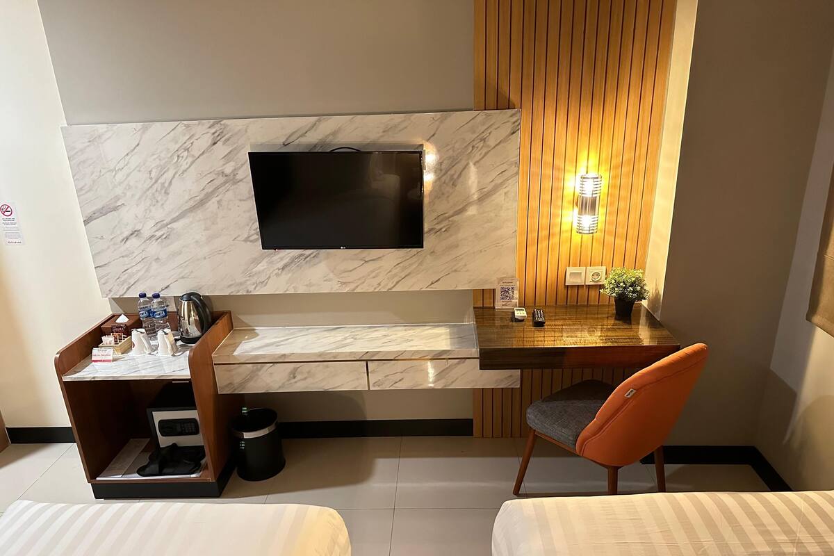 Ein Hotelzimmer mit Flachbildfernseher an einer Marmorwand, einem Holztisch mit einer kleinen Pflanze, einem Sessel und zwei Betten.