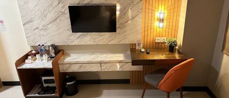 A hotel room with a flat-screen TV mounted on a marble wall, a wooden desk with a small plant, a chair, and two beds.