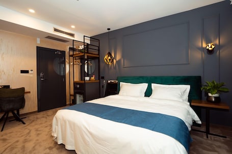 A modern hotel room with a large bed, a desk, a chair, and a small table with a plant.