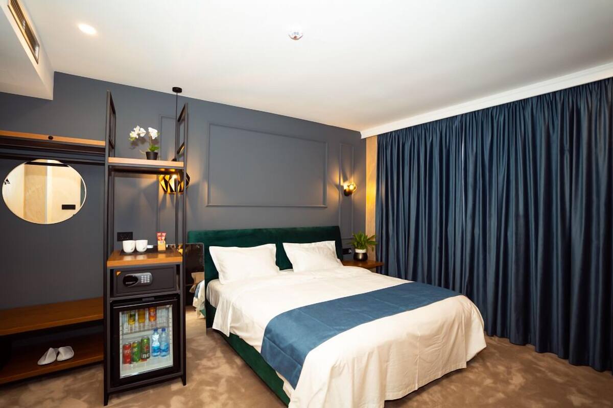 Une chambre d’hôtel avec un lit, un petit réfrigérateur, une table de chevet avec une lampe, un miroir et un rideau foncé.