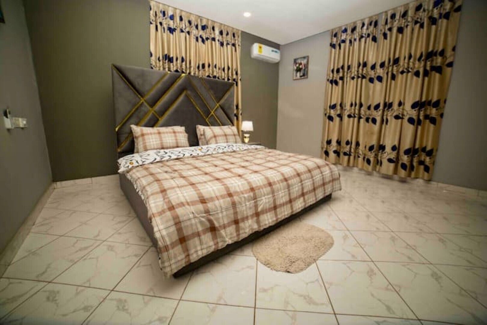 A bedroom with a bed, curtains, an air conditioning unit, and a tiled floor.