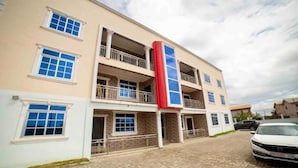A multi-story building with balconies, a central red column, and a paved driveway. - 3 bedrooms | Entire Flat | Amasaman, Accra (Amasaman,)