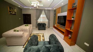 A modern living room with a sofa, a black armchair, a TV, and a wooden shelf. - 3 bedrooms | Entire Flat | Amasaman, Accra (Amasaman,)