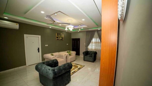 A spacious living area with a large dark sofa, a light-colored sofa, and a small armchair. The room has a tiled floor, a ceiling fan, recessed lighting, and a door leading to another room. - 3 bedrooms | Entire Flat | Amasaman, Accra (Amasaman,)