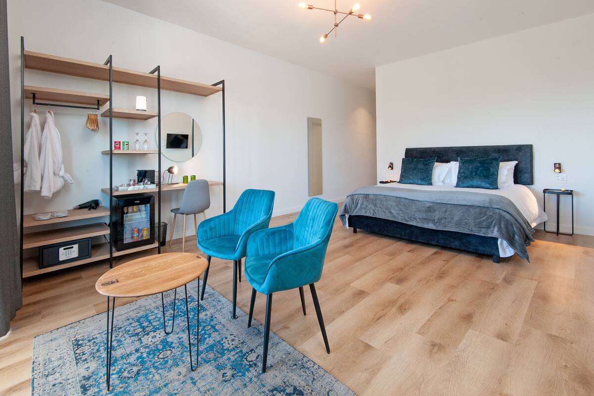 A modern hotel room with a bed, two blue chairs, a desk with a computer, a mirror, and a wardrobe.