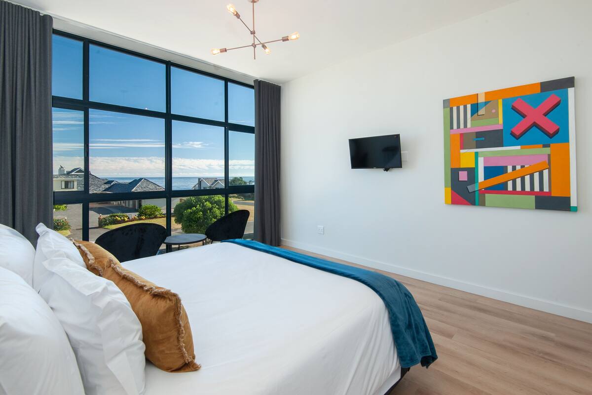 A hotel room with a large bed, a TV, a colorful abstract painting, and a view of the sea through large windows.