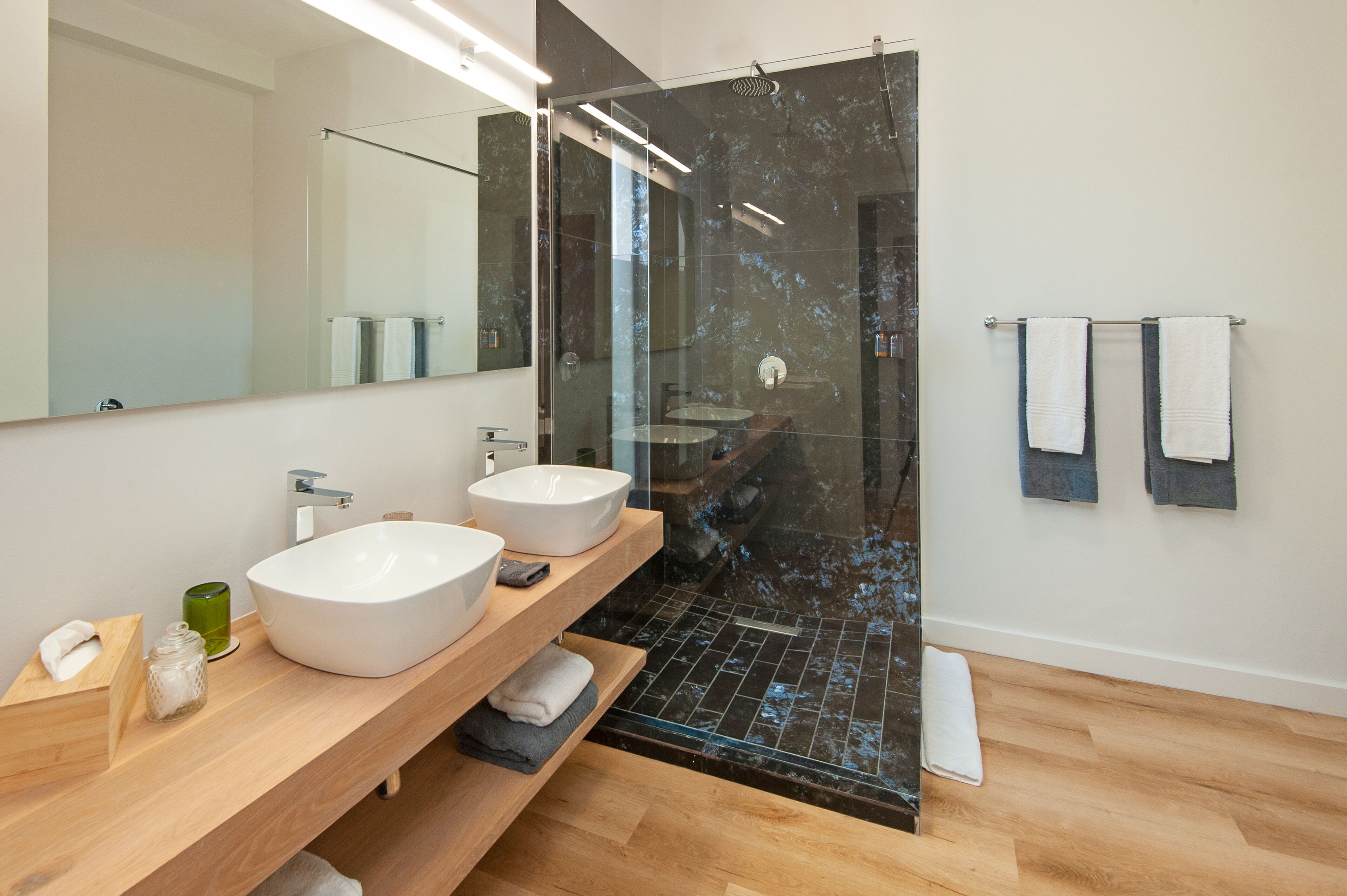 Un baño moderno con dos lavamanos blancos, un espejo grande y una ducha con cerramiento de vidrio.