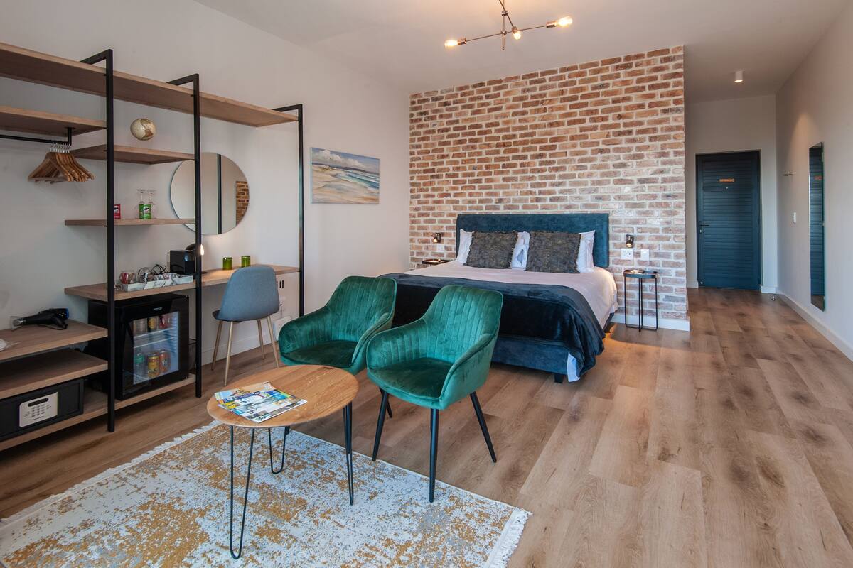 A modern bedroom with a brick accent wall, a large bed, green chairs, a wooden desk, and a small table.